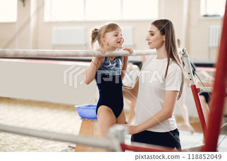 Child gymnastics balance beam. Girl gymnast athlete during an exercise horizontal bar in gymnastics competitions. Coach with child. 118852049