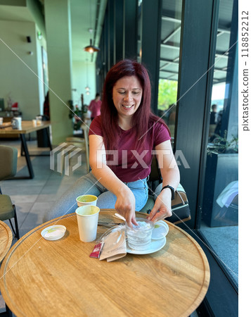 Woman with long red hair enjoys a casual moment at a coffee shop. Girl drink coffee in cafe. Bright interior and large windows create a cozy and inviting atmosphere. Breakfast time Woman with long red hair enjoys a casual moment at a coffee shop. Girl drink coffee in cafe. Bright interior and large windows create a cozy and inviting atmosphere. Breakfast time 118852212