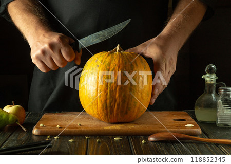A chef in a black uniform tries to cut a ripe pumpkin with a knife on a wooden board on a kitchen table. Asian national cuisine A chef in a black uniform tries to cut a ripe pumpkin with a knife on a wooden board on a kitchen table. Asian national cuisine 118852345