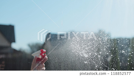 Woman in rubber gloves sprays window cleaner from a spray bottle onto the windows glass. Housewife hands in rubber gloves cleaning windows close up. Cleaning glass window with a cloth special rag blue 118852577