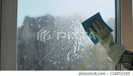 Woman in rubber gloves sprays window cleaner from a spray bottle onto the windows glass. Housewife hands in rubber gloves cleaning windows close up. Cleaning glass window with a cloth special rag blue 118852589