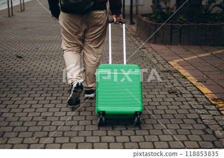 Unrecognizable man pulling green suitcase outdoors. Rear view. Copy space. Man walking on the street with travel bag. 118853835