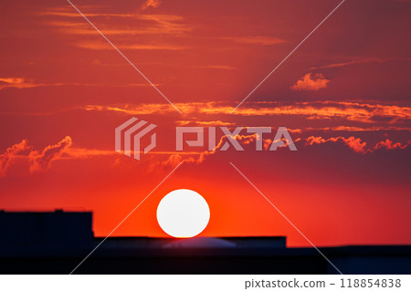 Stunning fiery sunset scene with vibrant red sky glowing sun setting behind city buildings urban silhouettes, creating warm dramatic atmosphere.  Tranquil horizon dusk cloudscape 118854838