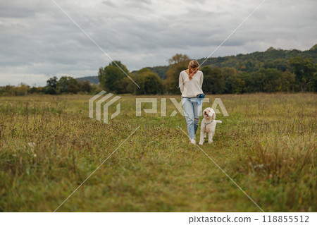 A Woman Taking a Leisurely Walk with Her Dog in a Scenic Field Beneath a Cloudy Sky 118855512