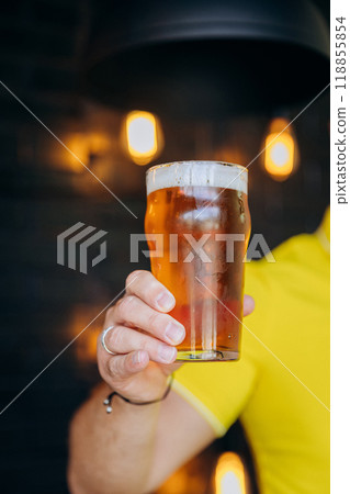 Close-up of person holding glass of beer with blurred pub background. Beer-related content 118855854
