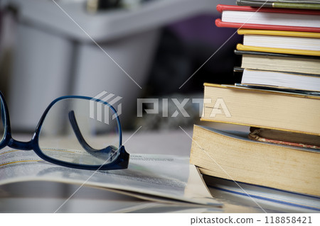 Reading glasses placed on an open book next to a stack of books, symbolizing a study or educational 118858421