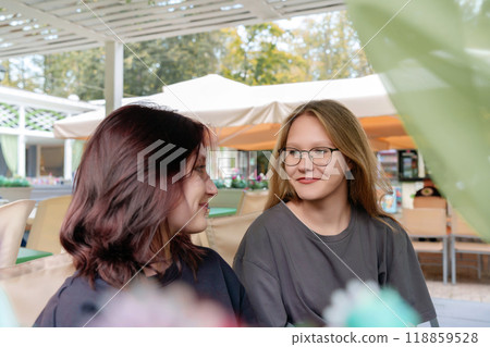 Cheerful female companions casually chatting and sharing a lighthearted moment while waiting for their order at a cozy summer coffee shop, capturing the fun and carefree spirit of friendship 118859528