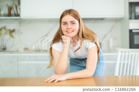 Portrait of positive teenage girl smiling at camera while leaning on countertop in cozy kitchen Portrait of positive teenage girl smiling at camera while leaning on countertop in cozy kitchen 118862266