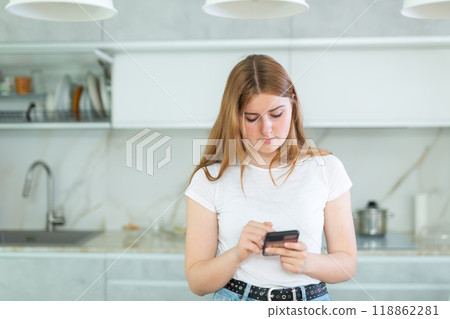 bored young woman stands in kitchen leans on countertop and surfs internet.Home pastime bored young woman stands in kitchen leans on countertop and surfs internet.Home pastime 118862281