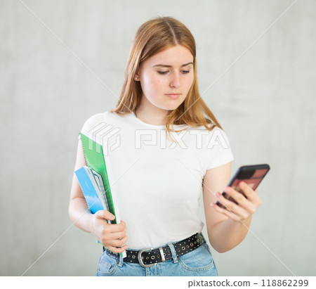 Positive teenage girl, student in casual clothes using mobile phone and holding spiral notebooks against gray wall background, copy space for mock up 118862299