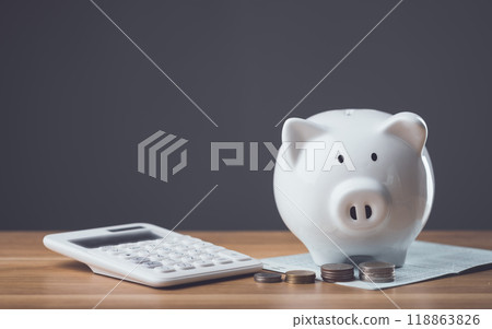 White piggy bank, bank book, and a pile of coins rest on a wooden table against a dark gray background. The scene symbolizes financial planning, savings, and the importance of managing money wisely. White piggy bank, bank book, and a pile of coins rest on a wooden table against a dark gray background. The scene symbolizes financial planning, savings, and the importance of managing money wisely. 118863826