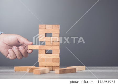 Businessman contemplates his next move in a stacking tower block game on a table against a gray wall background. Symbolizes strategic thinking, planning, and decision-making in business and life Businessman contemplates his next move in a stacking tower block game on a table against a gray wall background. Symbolizes strategic thinking, planning, and decision-making in business and life 118863971