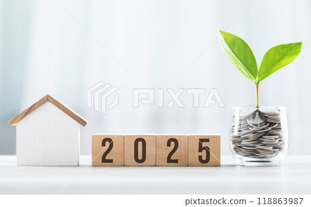 2025 is write on a wooden block next to glass jar filled with coins and a house model, placed on a wooden table against a bright white. Financial planning, savings, and effective money management 118863987
