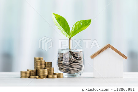 Glass jar filled with coins and a small seedling in the center, with stacked coins arranged from smallest to largest and a wooden house model on a table. Saving for house and financial growth 118863991
