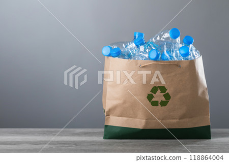 Concept of recycling is represented by old water bottles and clear plastic scraps placed in a paper bag on a table against a gray background. Reusing waste and promoting environmental sustainability 118864004