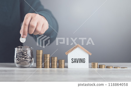 Man drops coins into a glass jar, with stacks of coins and a wooden house model beside him. Emphasizes the concept of saving money for future house investments and financial stability Man drops coins into a glass jar, with stacks of coins and a wooden house model beside him. Emphasizes the concept of saving money for future house investments and financial stability 118864058