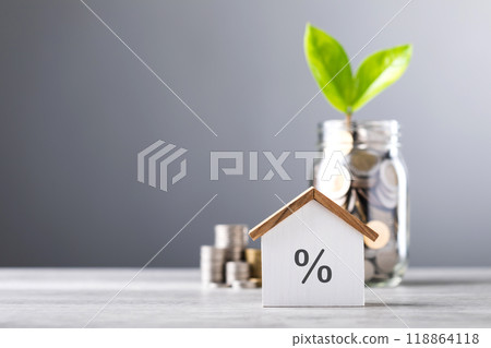 Glass jar filled with coins and a small seedling in the center, with stacked coins arranged from smallest to largest and a wooden house model on a table. Saving for house and financial growth Glass jar filled with coins and a small seedling in the center, with stacked coins arranged from smallest to largest and a wooden house model on a table. Saving for house and financial growth 118864118