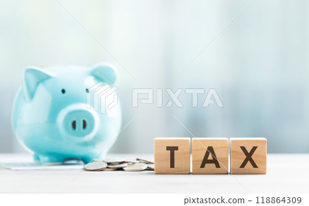 TAX is written on a wooden block beside a blue piggy bank and a pile of coins on a wooden table against a bright white background. Financial planning, savings, and wise money management. TAX is written on a wooden block beside a blue piggy bank and a pile of coins on a wooden table against a bright white background. Financial planning, savings, and wise money management. 118864309
