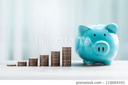 A blue piggy bank and stack of coins are neatly arranged on a table against a bright white background. The image represents the concept of saving money for the future. A blue piggy bank and stack of coins are neatly arranged on a table against a bright white background. The image represents the concept of saving money for the future. 118864341