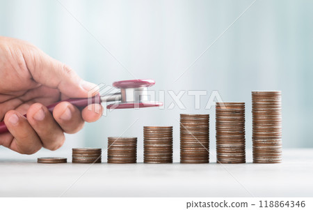 Hand holding a stethoscope, examining a stack of coins on a table against a bright white background. The image symbolizes the concept of a financial health check and monetary well-being. 118864346