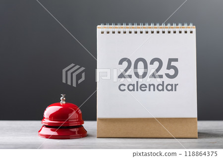 Red service bell and a 2025 desk calendar are placed on a table against a gray background. The image represents the concept of time management, planning, and readiness for the upcoming year. 118864375
