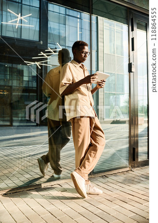 Vertical long shot of young Black office worker standing outdoors scrolling news feed in social network on digital tablet Vertical long shot of young Black office worker standing outdoors scrolling news feed in social network on digital tablet 118864555
