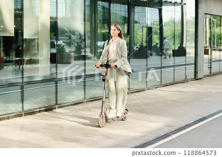 Wide shot of young Caucasian woman wearing stylish formal clothes riding e-scooter along business district, copy space 118864573
