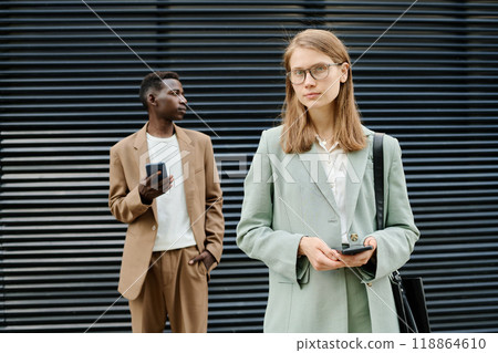 Medium portrait of young Caucasian female manager holding smartphone and looking at camera, her Black male colleague standing relaxed in background 118864610