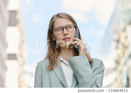 Low angle medium closeup of young Caucasian female manager wearing eyeglasses having phone call while walking outdoors Low angle medium closeup of young Caucasian female manager wearing eyeglasses having phone call while walking outdoors 118864612