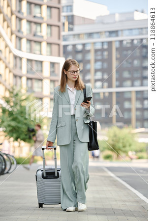 Young Caucasian woman on business trip texting on smartphone while walking along city street with her luggage Young Caucasian woman on business trip texting on smartphone while walking along city street with her luggage 118864613