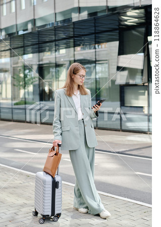 Young female manager wearing stylish outfit standing outdoors with her luggage and scrolling news feed on smartphone while waiting for taxi 118864628