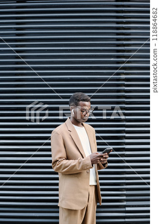 Vertical medium long shot of young Black manager wearing stylish outfit standing outdoors in business district using smartphone 118864682