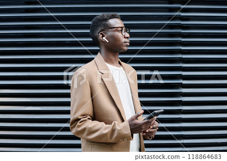 Medium shot of young African American businessman holding smartphone listening to voice message via earphones while standing outdoors 118864683