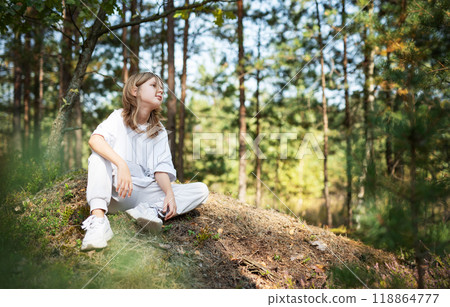 Young girl enjoying a sunny day in a forest clearing while sitting on a grassy mound Young girl enjoying a sunny day in a forest clearing while sitting on a grassy mound 118864777