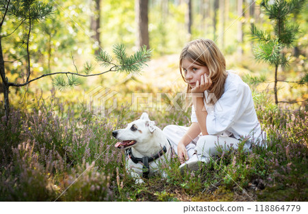 A young girl sitting among wildflowers in a forest, gently resting her chin on her hand with her dog 118864779