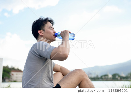 A tired, sporty Asian man in sportswear sits by the lake, drinking to refresh himself after a run. 118864901