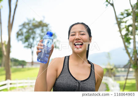 A fit Asian woman in sportswear is holding up her water bottle, standing in a green park. 118864903