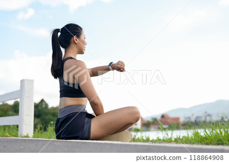 A sporty Asian girl in sportswear sits by the lake, checking her running miles on her smartwatch. A sporty Asian girl in sportswear sits by the lake, checking her running miles on her smartwatch. 118864908