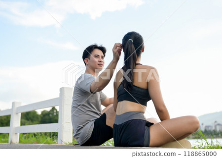 A lovely, sporty Asian couple in sportswear sits by the lake, resting after a morning run together. A lovely, sporty Asian couple in sportswear sits by the lake, resting after a morning run together. 118865000