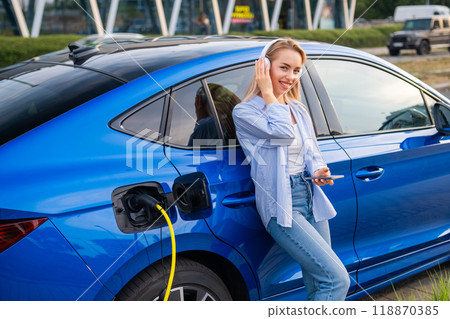 Woman monitors her electric cars charge status using her smartphone, while wearing headphones standing near the blue car. Woman monitors her electric cars charge status using her smartphone, while wearing headphones standing near the blue car. 118870385