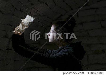 Young adult in a basement looking in a small window. A beam of light falls on her face. Portrait of the young girl in the dungeon with dark background. Young adult in a basement looking in a small window. A beam of light falls on her face. Portrait of the young girl in the dungeon with dark background. 118877480