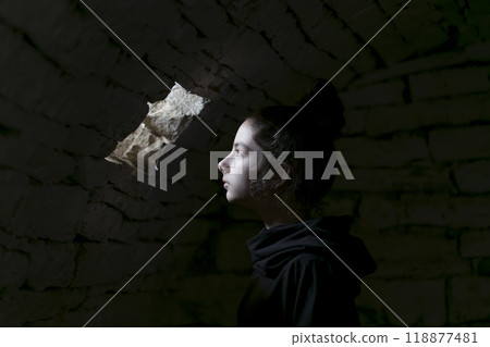 Young adult in a basement looking in a small window. A beam of light falls on her face. Portrait of the young girl in the dungeon with dark background. Young adult in a basement looking in a small window. A beam of light falls on her face. Portrait of the young girl in the dungeon with dark background. 118877481