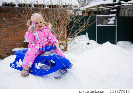 Sitting on a sled on a snowy hill, a child girl in a bright pink jumpsuit happily waves at the camera. Copy Space. A child happily sledding down an icy hill on a wonderful winter day. Sitting on a sled on a snowy hill, a child girl in a bright pink jumpsuit happily waves at the camera. Copy Space. A child happily sledding down an icy hill on a wonderful winter day. 118877613