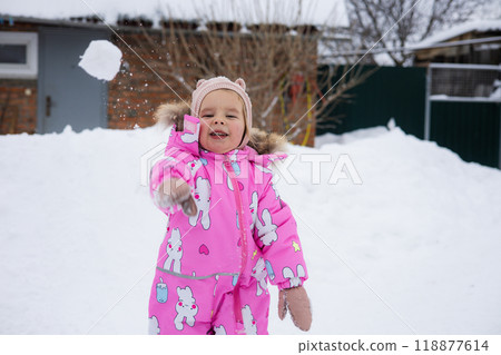 Happy baby in pink snowsuit playing in snow, smiling and throwing snowballs on cold winter day. Cute child laughing happily and throwing snowball at camera on winter day in yard. Happy baby in pink snowsuit playing in snow, smiling and throwing snowballs on cold winter day. Cute child laughing happily and throwing snowball at camera on winter day in yard. 118877614
