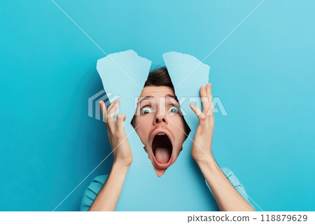 A young man expressing shock and surprise through a torn blue wall effect in a studio setting A young man expressing shock and surprise through a torn blue wall effect in a studio setting 118879629
