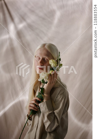 Vertical shot of young Caucasian woman with albinism holding white lisianthus flowers standing in studio decorated with beige fabrics 118883554