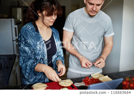 A young couple prepares Italian sandwiches following a video recipe on a tablet. 118883684