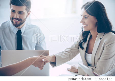 So nice to meet you! Close-up part of young beautiful women shaking hands with smile while sitting at the office table with her coworkers 118884178