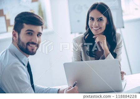 Enjoying their work together. Two cheerful coworkers looking at camera with smile while sitting at the office table 118884228