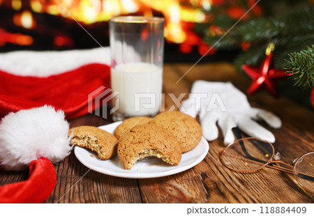 a bitten oatmeal cookie and an unfinished glass of milk on a wooden table against the background of Santa Claus clothes, a Christmas tree and a fireplace 118884409